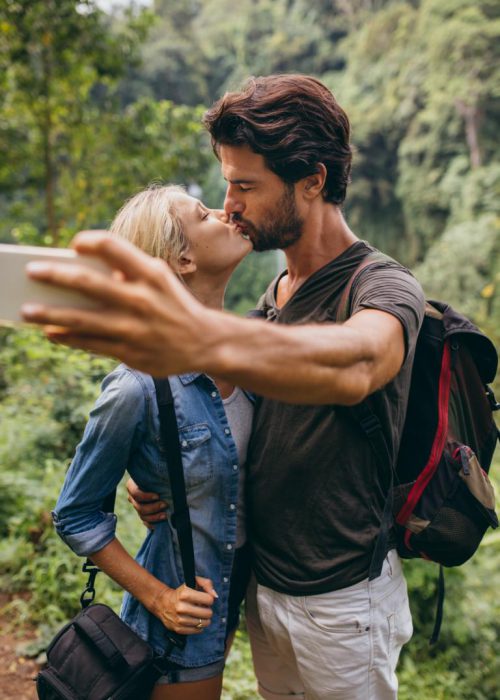 Loving young couple kissing and taking self portrait with their cell phone. Couple in love taking self picture in forest.
